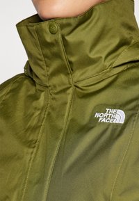 Gros plan d'une personne portant une veste North Face vert olive avec le col fermé et le logo visible sur la poitrine.