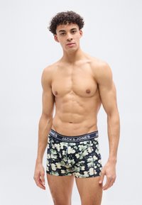 Νεαρός άνδρας με σγουρά μαλλιά, φοράει μαύρα ανδρικά εσώρουχα boxer με λουλουδάτο μοτίβο Jack & Jones, στέκεται μπροστά σε απλό ανοιχτό γκρι φόντο.