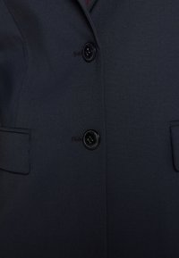 Blazer bleu marine en tissu texturé, avec deux boutons noirs, deux poches avant et un design de revers subtil.