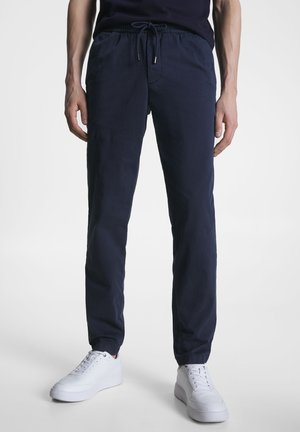 Hombre con pantalones jogger azul marino con puños elásticos y zapatillas blancas, de pie frente a un fondo claro y liso.