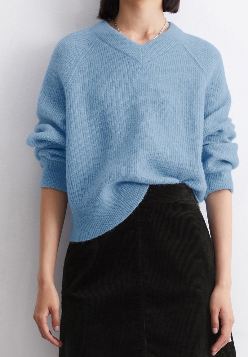 Personne portant un pull tricoté côtelé bleu clair à manches longues et une jupe noire taille haute sur un fond blanc uni.