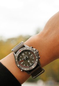 Reloj analógico gris con esfera negra, marcadores numéricos, acentos rojos y correa de tela gris, mostrado en una muñeca.