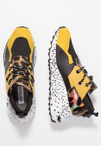 Les baskets noires et jaunes présentent une tige texturée avec des accents imprimé léopard et une semelle blanche tachetée, détaillée avec des lacets noirs.