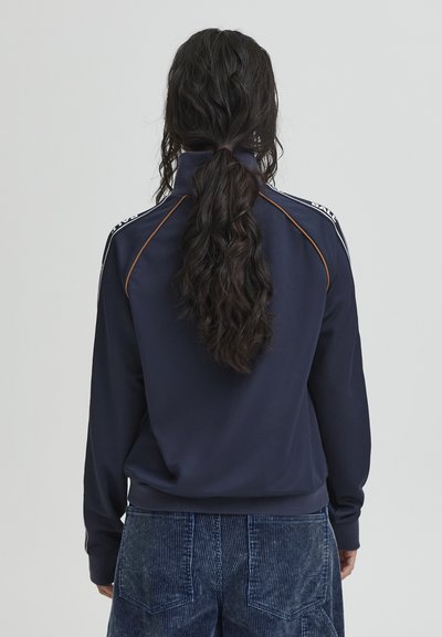 Granatowa bluza z wysokim kołnierzem, kontrastującym pomarańczowym przeszyciem oraz białymi paskami na ramionach, zestawiona ze spodniami sztruksowymi.