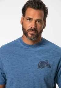 Modré tričko s krátkým rukávem vyrobené z texturované látky, s černým logem "The Great Industrial" na levé části hrudi.