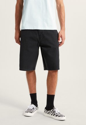 Czarne szorty dżinsowe o luźnym kroju, do kolan, w zestawieniu z czarnymi skarpetkami i trampkami w zebra. Prosty design, bez widocznych logo.