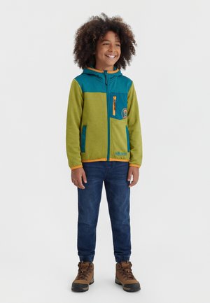 Usmievavé dieťa s kučeravými vlasmi, oblečené v zelenej a tyrkysovej mikine na zips, tmavomodrých džínsoch a hnedých turistických topánkach, stojace na bielom pozadí.