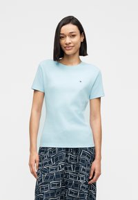 Νεαρή γυναίκα με ίσια σκούρα μαλλιά φοράει ανοιχτό μπλε εφαρμοστό μπλουζάκι και σκούρο μπλε παντελόνι με σχέδια, στέκεται μπροστά σε λευκό φόντο.