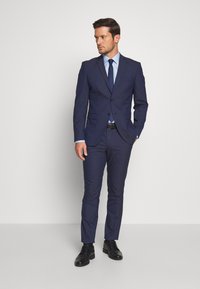 Marinblå kostym med smal passform, som har enblads blazer, matchande byxor och en ljusblå skjorta. Svarta dressade skor fulländar outfiten.