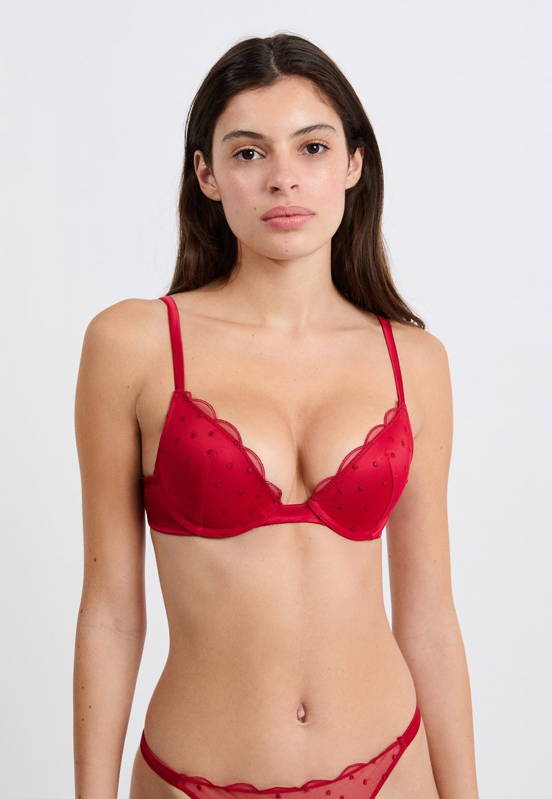 Mujer con cabello oscuro y largo que lleva puesta una braga de encaje rojo y ropa interior a juego sobre un fondo blanco liso.