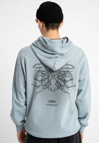 Jasnoniebieska bluza z kapturem z dużym czarnym wzorem na plecach. Prezentuje luźny krój, długie rękawy oraz przestronny kaptur z zapięciem na napy.