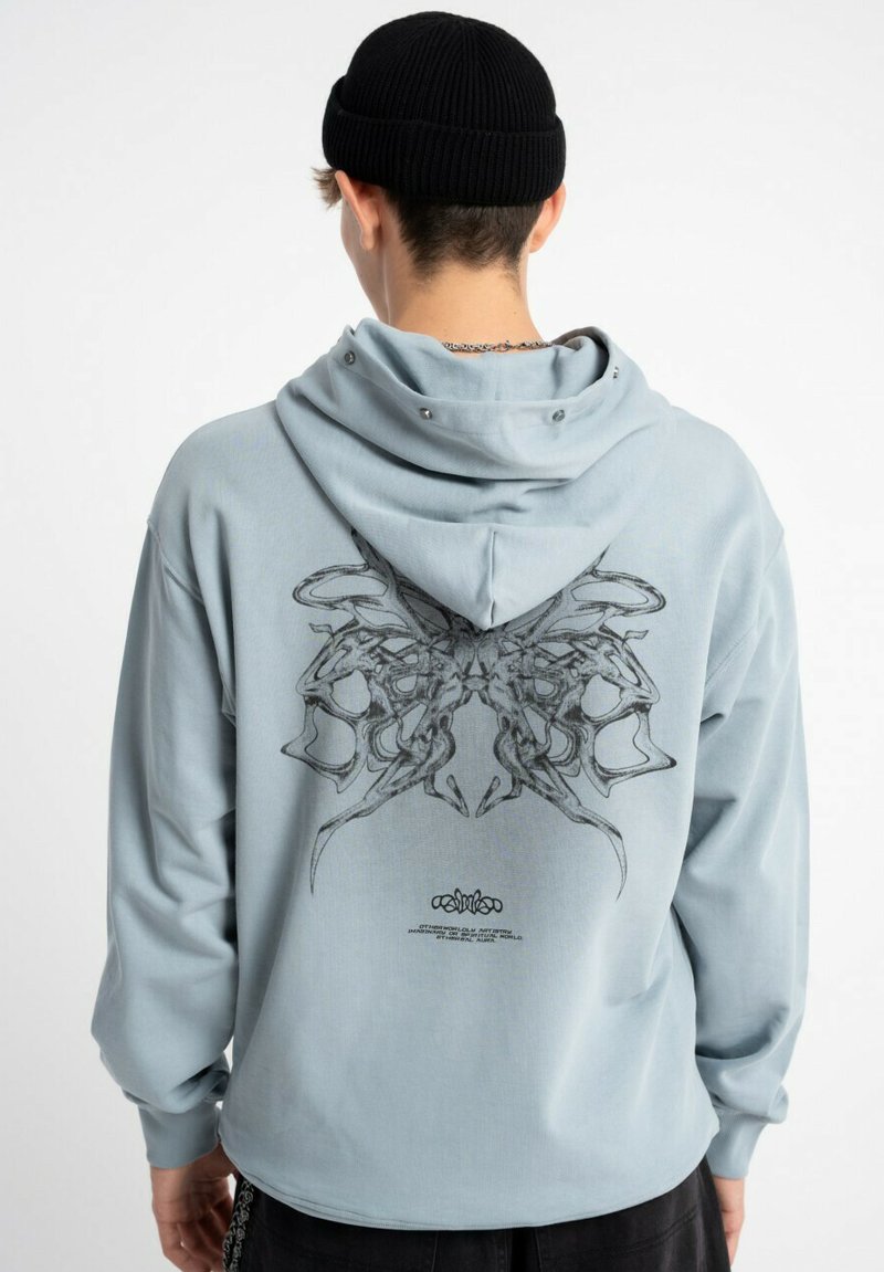 Jasnoniebieska bluza z kapturem z dużym czarnym wzorem na plecach. Prezentuje luźny krój, długie rękawy oraz przestronny kaptur z zapięciem na napy.