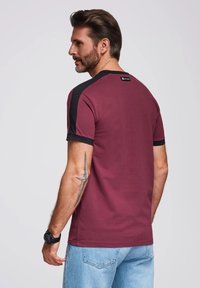 Bordeaux tričko s krátkým rukávem a černými akcenty na ramenech a rukávech. Vyrobeno z měkkého materiálu, má štítek blízko výstřihu.