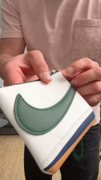 Λευκό αθλητικό παπούτσι με πράσινη δερμάτινη λωρίδα, ανάγλυφη επιφάνεια, αντίθετη σόλα σε ναυτικό και καουτσούκ γκας, και διακοσμητική λεπτομέρεια με φερμουάρ.