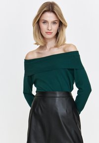 Sweter w kolorze ciemnozielonym z dekoltem typu off-the-shoulder, prążkowany, połączony z czarną skórzaną spódnicą o wysokim stanie. Gładka faktura, dopasowany krój i bezszwowe akcenty.