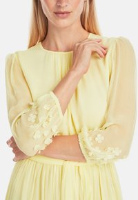 Gelbes, plissiertes Kleid mit durchsichtigen, puffärmeligen Ärmel, verziert mit floralen Applikationen und einem lässigen Schnitt, mit einem runden Ausschnitt und passendem Gürtel.