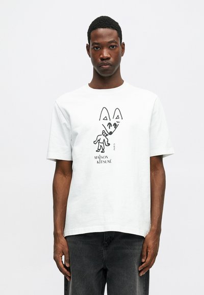 Muž v bílém tričku s černým abstraktním grafickým motivem liščí tváře a postavy, s nápisem "Maison Kitsuné Paris," doplněný tmavými džíny.