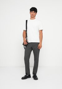 Witte katoenen t-shirt, grijs geruite broek, zwarte schoenen. Model staat met camerariem over de schouder, op een effen achtergrond.