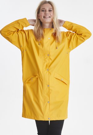 Femme souriante portant un long imperméable jaune avec boutons-pression et poches, les mains derrière la tête, debout devant un fond uni.
