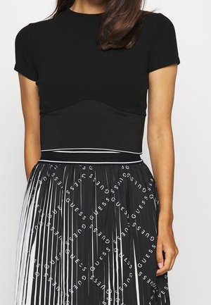 Mujer vistiendo una blusa negra de manga corta y una falda negra plisada con rayas verticales blancas y un patrón repetido del logo "GUESS".
