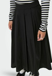Gonna nera plissettata con una trama liscia, vita alta, abbinata a scarpe nere in vernice e a una maglietta a maniche lunghe a righe bianche.