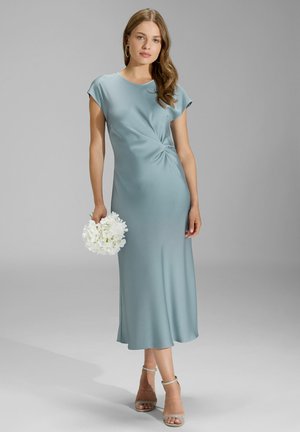 Maxi sukienka w miękkim niebieskim materiale, z krótkimi rękawami w formie bufek, skręconym detalem w talii i zwiewnym dołem. Modelka trzyma białe kwiaty.