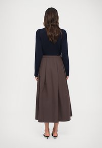 Vrouw met lang bruin haar, gekleed in een marineblauwe trui, een bruine midi rok en zwarte hakken, staand met haar rug naar de camera.