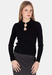 Donna bionda che indossa un top nero a maniche lunghe a costine con ritagli decorativi e bottoni, abbinato a una gonna grigia, in piedi davanti a uno sfondo bianco.