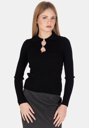 Donna bionda che indossa un top nero a maniche lunghe a costine con ritagli decorativi e bottoni, abbinato a una gonna grigia, in piedi davanti a uno sfondo bianco.