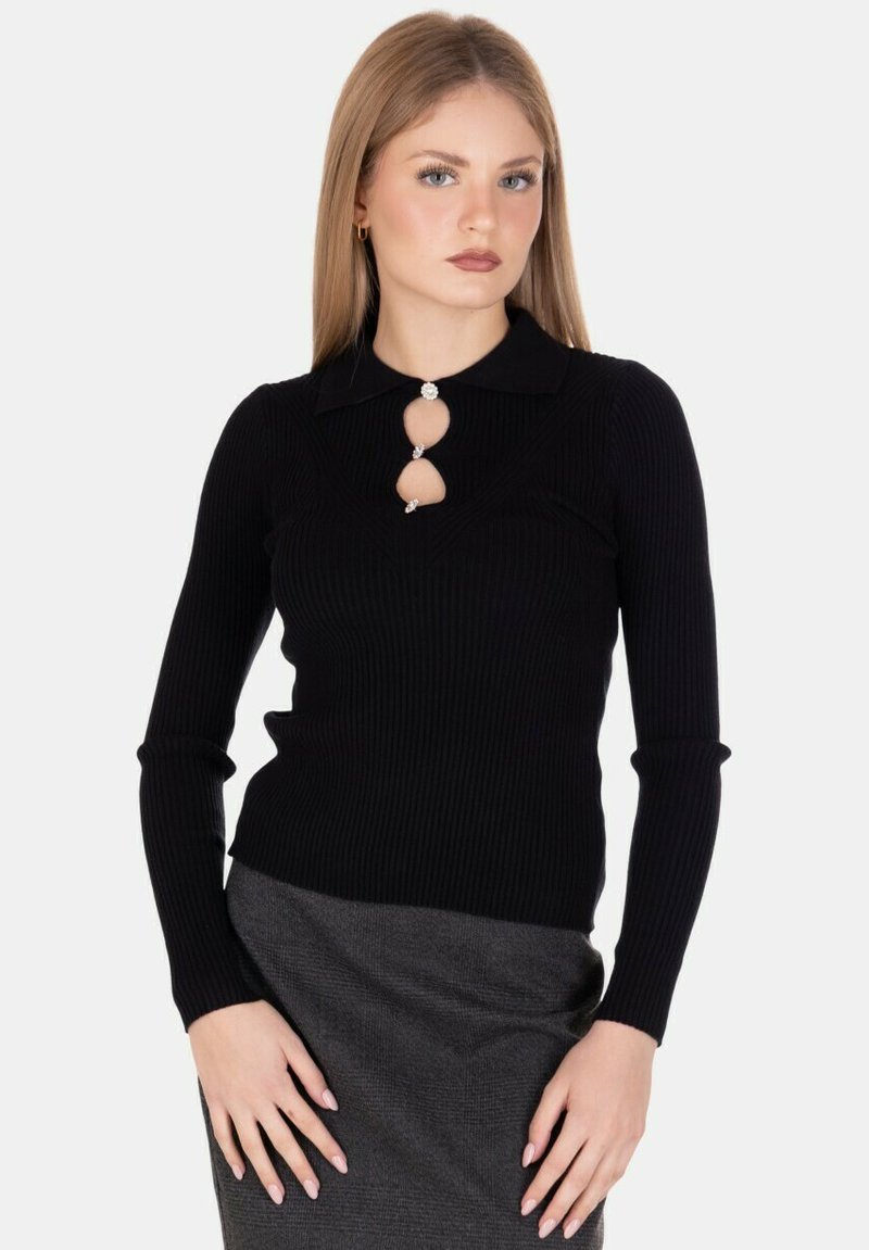 Donna bionda che indossa un top nero a maniche lunghe a costine con ritagli decorativi e bottoni, abbinato a una gonna grigia, in piedi davanti a uno sfondo bianco.