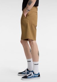Shorts en coton couleur fauve avec poches latérales, dotés d'une coupe droite et d'ourlets, portés avec des baskets bleues et noires et des chaussettes blanches.