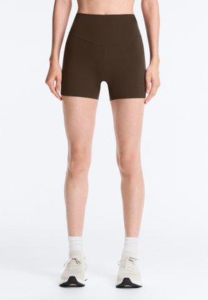 Kvinde iført brune, højtaljede, tætsiddende shorts, hvide knæstrømper og hvide træningssko, stående mod en ensfarvet baggrund.