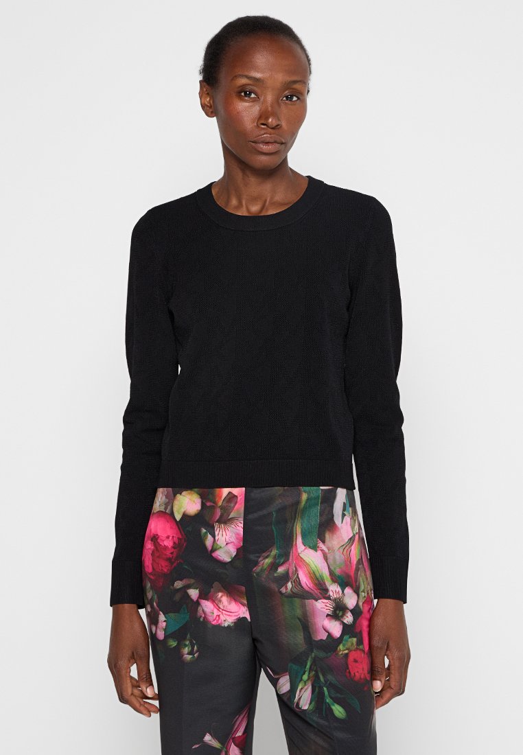 Donna che indossa un maglione nero a maniche lunghe e pantaloni neri con grandi motivi floreali rosa e verdi, in piedi di fronte a uno sfondo chiaro e semplice.