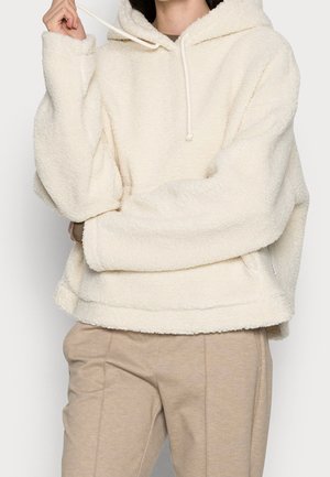 Personne portant un sweat à capuche crème et duveteux avec des cordons et un pantalon beige, tenant un cordon près du visage.