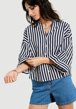 Frau, die den Ärmel eines locker sitzenden, schwarz-weiß gestreiften Hemds mit Stehkragen hochkrempelt, trägt blaue Jeansshorts, ein goldenes Armband und eine Halskette.