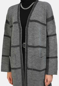 Cardigan grigio con motivo geometrico nero, apertura frontale, maniche lunghe e due tasche anteriori, realizzato in tessuto lavorato a maglia. Sotto, una semplice maglietta nera.