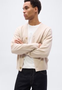 Νεαρός άνδρας με σταυρωμένα χέρια, φορούσε ένα μπεζ ζακέτα πάνω από ένα άσπρο πουκάμισο και σκούρα τζιν, κοιτάζοντας προς τα πλάγια μπροστά σε απλό φόντο.