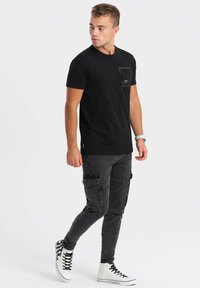 T-shirt nera in cotone con tasca sul petto, abbinata a pantaloni cargo grigi slim-fit con più tasche e sneakers bianche alte.
