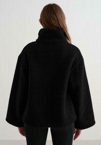 Puhaszőrű fekete fleece pulóver magas gallérral és széles ujjakkal. Az anyaga lágy, vastag és kényelmes, ideális hűvösebb időjárásra.