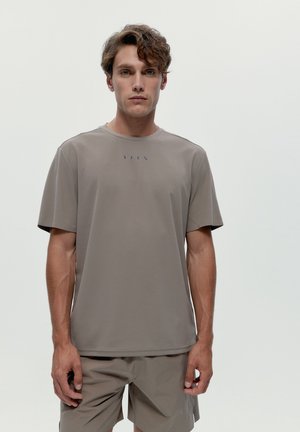Junger Mann mit welligem braunem Haar, der ein taupefarbenes kurzärmeliges T-Shirt und passende Shorts trägt und vor einem schlichten hellen Hintergrund steht.