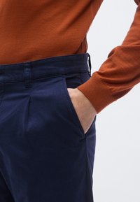 Marinblå byxor med texturerad yta, utrustade med sidofickor och en skräddarsydd passform, ihop med en rostfärgad figursydd tröja.