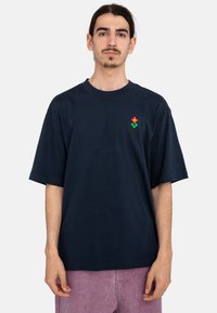 Camiseta de algodón azul marino con cuello redondo y mangas cortas, que presenta un pequeño logo de flor roja con acentos verdes en el pecho.