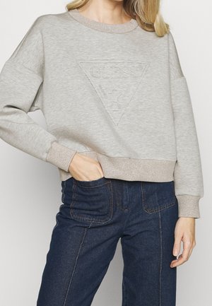 Kobieta w jasnoszarej bluzie z subtelnym tłoczonym logo Guess i ciemnoniebieskich dżinsach, z ręką w kieszeni, na jednolitym tle.