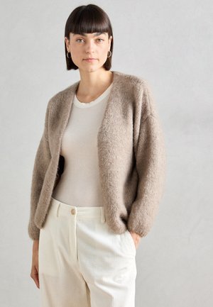 Gilet en tricot beige avec une texture douce, à l'avant ouvert et avec des manches légèrement surdimensionnées, porté sur une chemise ajustée de couleur claire et un pantalon en velours côtelé.