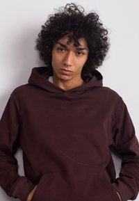 Felpa marrone con cappuccio, realizzata in un tessuto testurizzato, con tasca a marsupio e design minimalista. La vestibilità è comoda, adatta per un look casual.