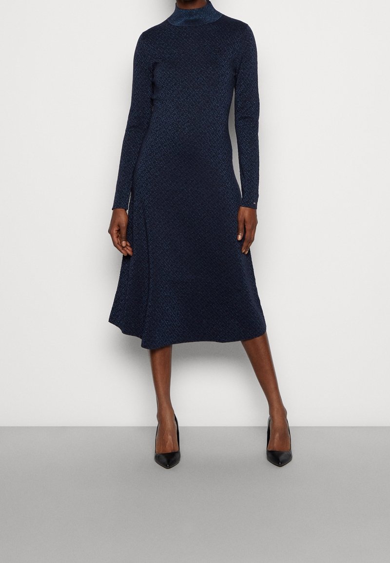 Robe bleu marine à manches longues avec un col montant, un ourlet à hauteur des genoux, un motif texturé et une couture avant en biais. Associée à des escarpins noirs à bout pointu.