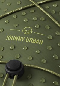 Grönt vattentåligt material med upphöjda prickar, "JOHNNY URBAN"-logotyp och en svart snörlås med matchande olivgrönt snöre.