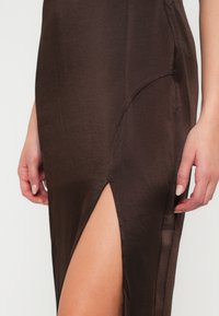 Hnedé saténové šaty s bočným rozparkom, ktoré majú hladkú textúru a priliehavý dizajn. Záber zblízka sa zameriava na látku a umiestnenie ruky.