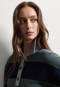 Suéter de polo con cuello gris, cuerpo verde y rayas azul marino. Cuenta con cuatro botones y un tejido texturizado. El tono de piel es claro.