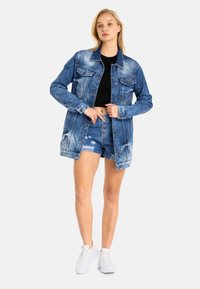 Jeansjack met een lichte wassing, twee borstzakken en knoopsluiting. Gecombineerd met versleten denim shorts en witte sneakers.